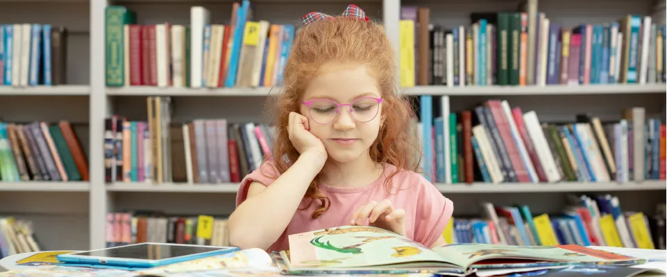 画像：本がたくさん並んでいる本棚があり、その前のテーブルに本やタブレットが置いてあり、メガネをかけた女の子が本を読んでいる様子。